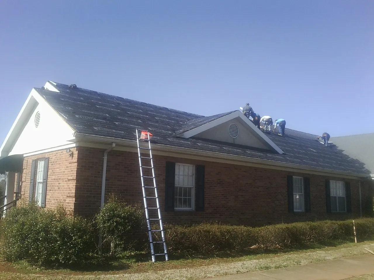 Crew tearing shingles off a brick ranch roof with a ladder set — Metal Roof Repair work by our crew in Black River