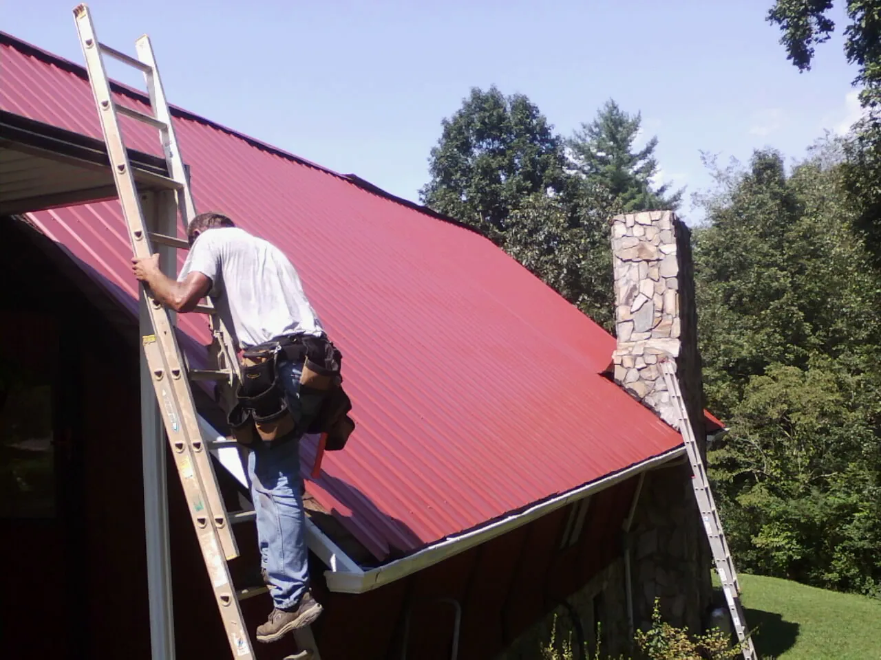 Roofer on a ladder beside a red metal roof and stone chimney — Soffit & Fascia Repair by Roofing Brothers in Black River